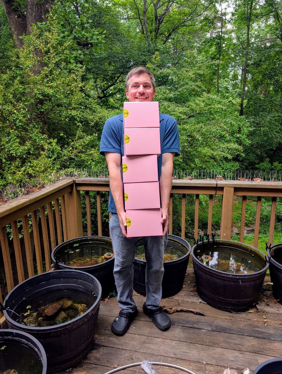 Chris holding a stack of Firefly Medaka shipping boxes on the deck, with the outdoor medaka tubs around him