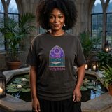 Woman with natural black curls wearing the Cathedral Pond tee in Graphite, in a stone conservatory with gothic windows and a lily pond