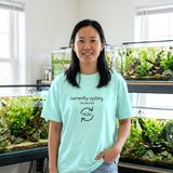 Woman with shoulder-length black hair wearing the Currently Cycling tee in Chalky Mint, in a bright fish room with planted aquariums on a metal rack