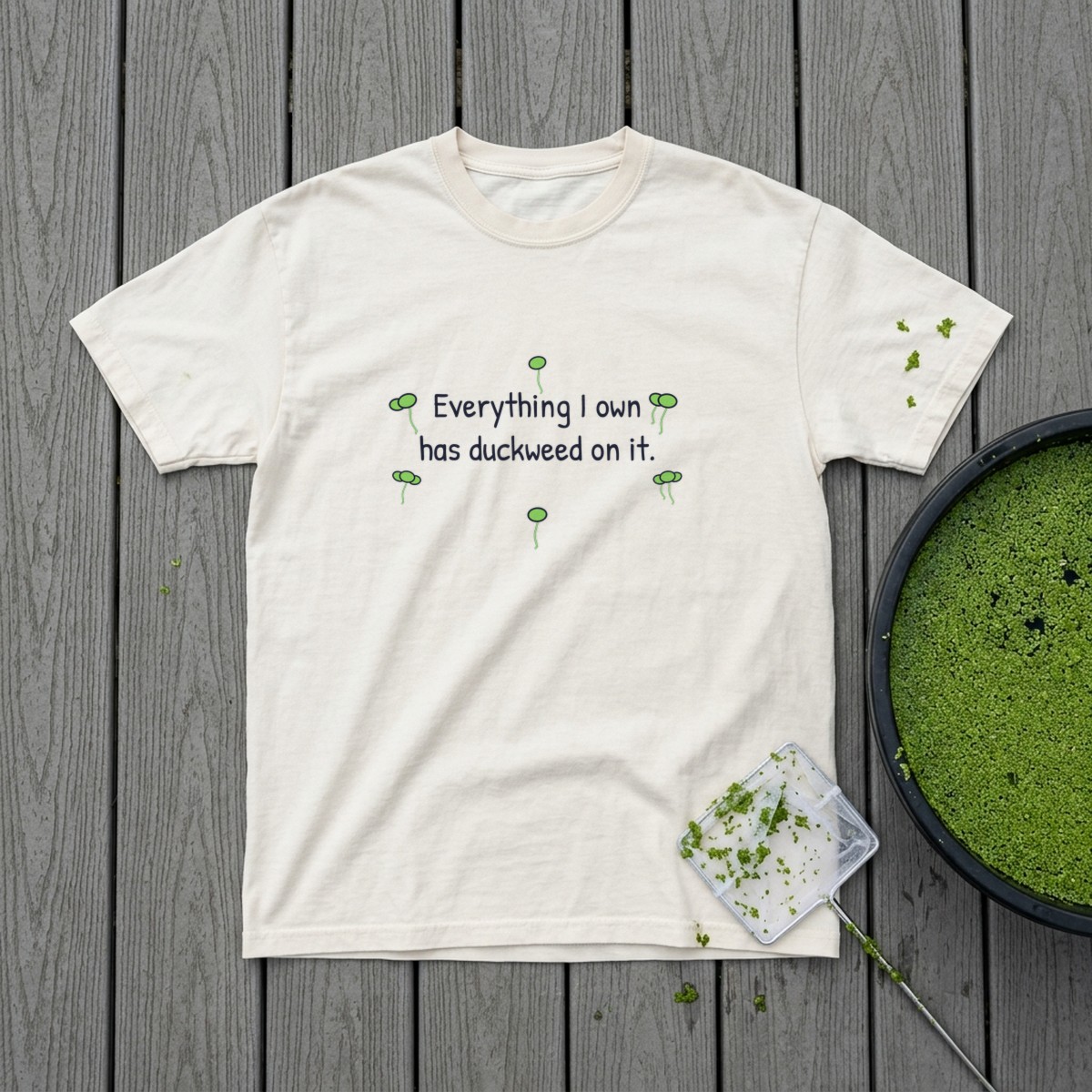 Everything I own has duckweed on it t-shirt in Ivory laid flat on gray deck boards beside a black tub completely covered in duckweed and a white net with duckweed stuck to it, a few stray fronds on the sleeve
