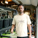Man with glasses and a short beard wearing the Everything I own has duckweed on it tee in Ivory, holding a dripping net of duckweed in a garage fish room