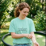 Woman with curly auburn hair wearing the Everything I own has duckweed on it tee in Chalky Mint, picking duckweed off her wrist beside a stock tank pond covered in duckweed