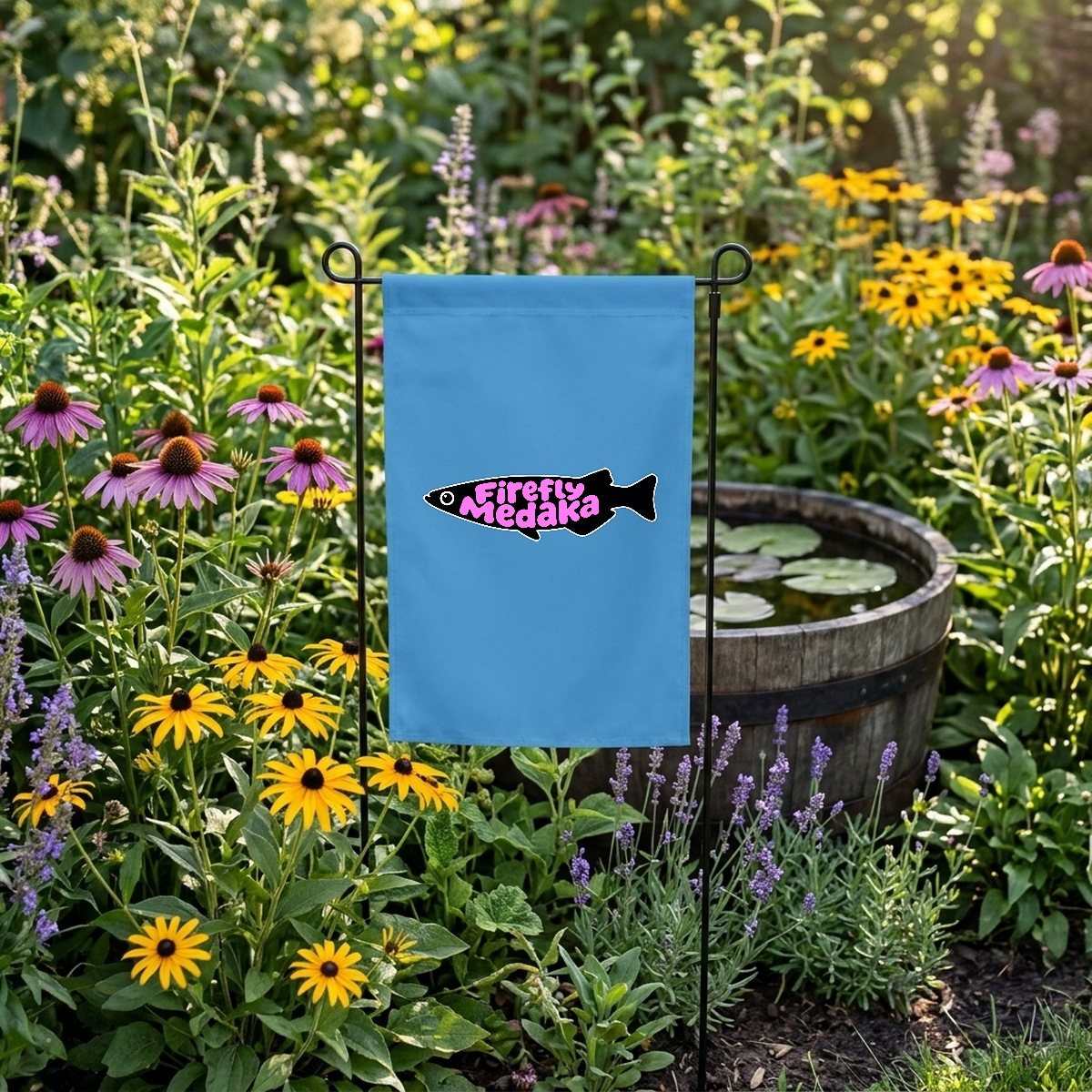 Firefly Medaka logo garden banner in sky blue on a black garden stake in a summer flower bed beside a half-barrel water lily tub