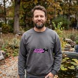 Man with a full beard wearing the Firefly Medaka logo crewneck sweatshirt in Charcoal, beside a stock tank pond in an autumn backyard