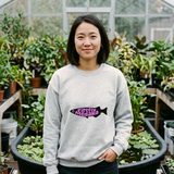 Woman with a dark bob wearing the Firefly Medaka logo crewneck sweatshirt in Sport Grey, in a greenhouse beside a tub pond of lilies and floating plants