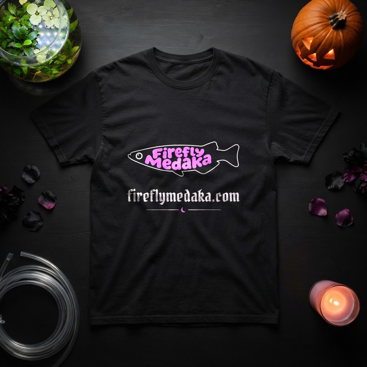 Firefly Medaka Nightshade logo t-shirt in black on a black table beside a glowing planted fishbowl, a jack-o'-lantern, aquarium airline tubing, petals, and a pink candle