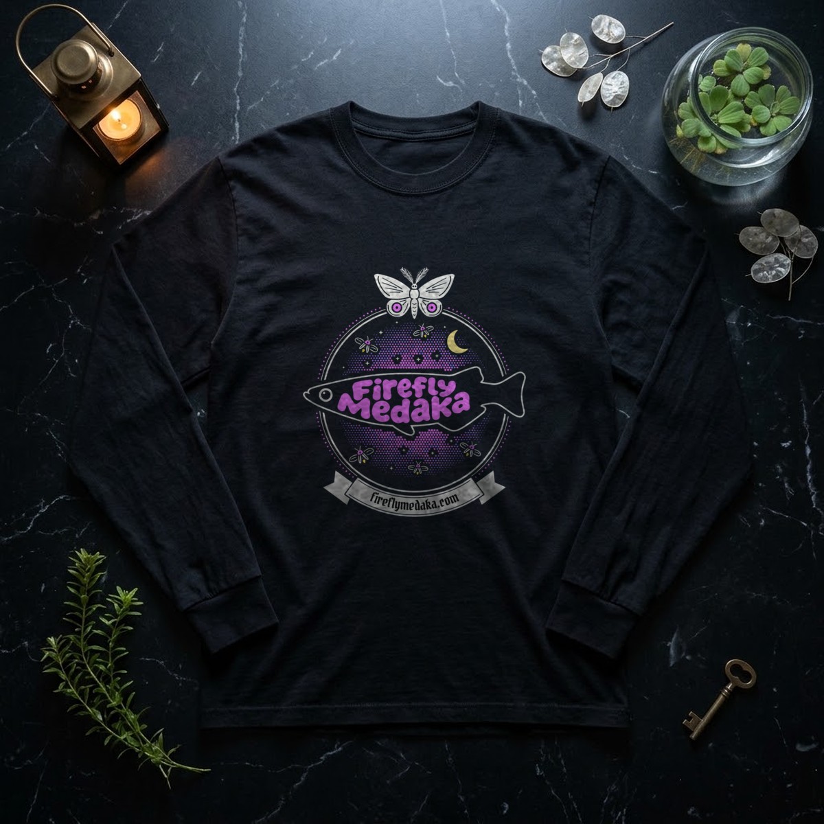 Firefly Medaka Nocturne logo long sleeve shirt in black on polished black marble beside a brass lantern, a glass jar of floating water lettuce, an aquarium stem plant, silver honesty seed pods, and an old brass key