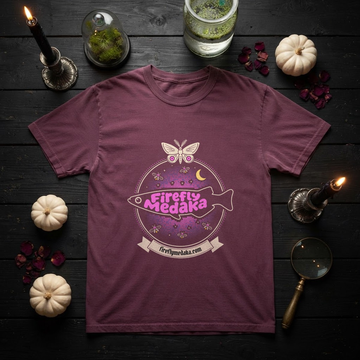 Firefly Medaka Nocturne logo t-shirt in Berry on black boards with white pumpkins, a moss cloche, a jar of duckweed, a magnifying glass, and black taper candles