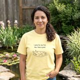 Woman with curly dark hair wearing the I Came for One Fish tee in Butter, at the stone edge of a backyard pond with blooming water lilies