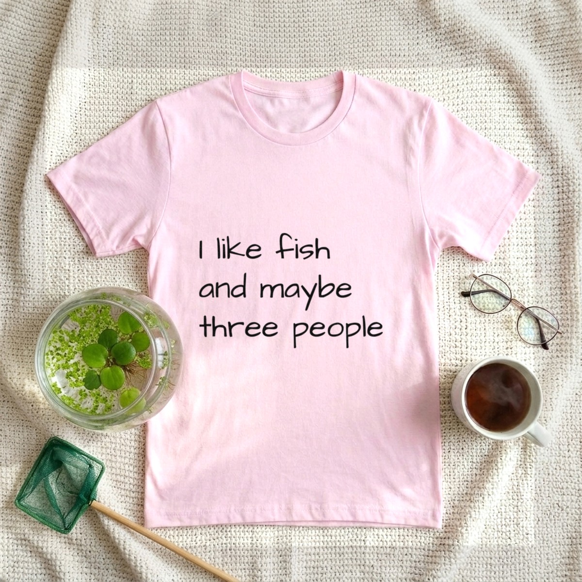 I like fish and maybe three people t-shirt in light pink, laid flat on a cream knit blanket with a glass bowl of duckweed and frogbit, a fish net, reading glasses, and tea