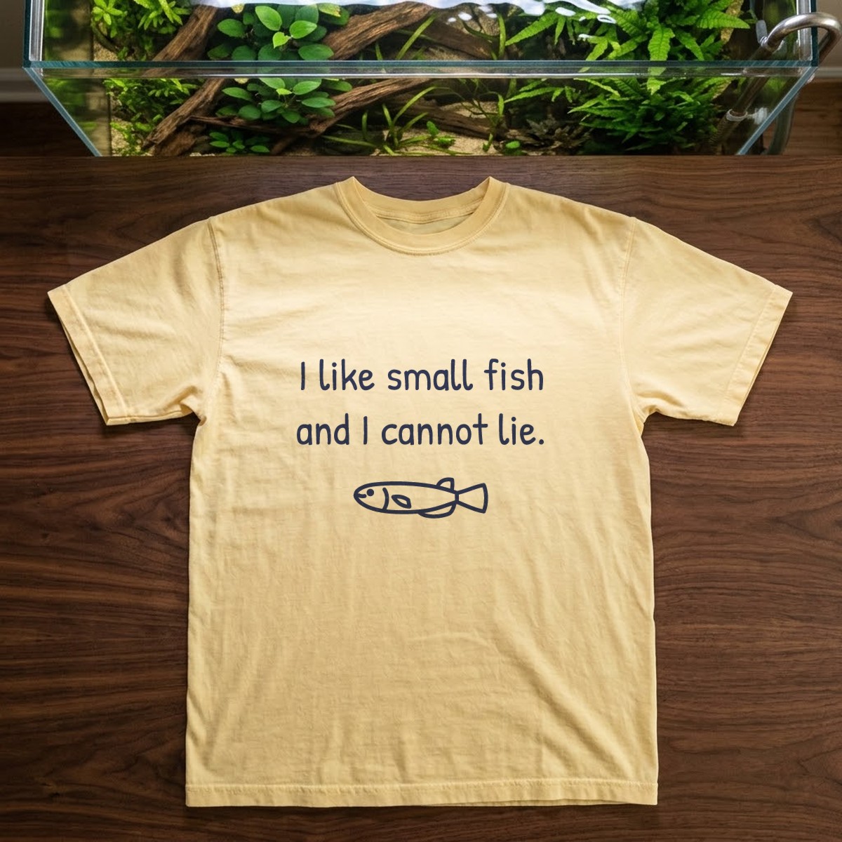 I like small fish and I cannot lie tee in butter yellow, laid flat on a dark walnut table beside the open top of a large planted aquarium