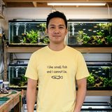 Man with short black hair wearing the I Like Small Fish and I Cannot Lie tee in Butter, in a home fish room with rows of planted breeder tanks