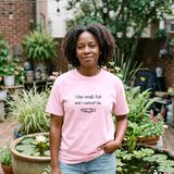 Woman with natural curly hair wearing the I Like Small Fish and I Cannot Lie tee in Blossom, on a brick patio with container ponds of water lilies