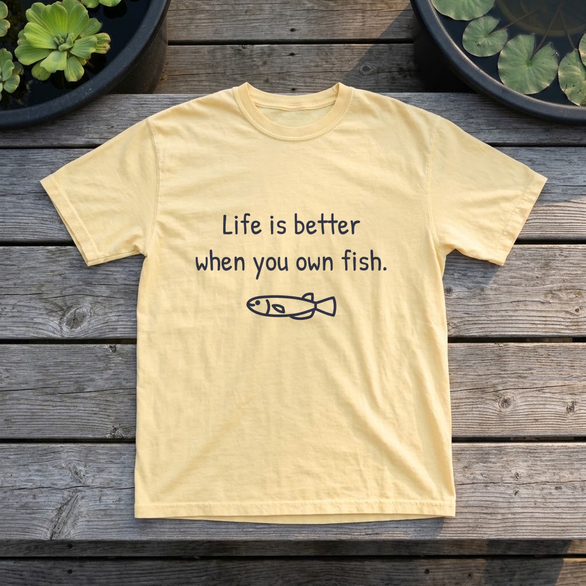 Life is better when you own fish tee in butter yellow, laid flat on weathered backyard bench boards between outdoor tubs of water lettuce and lily pads