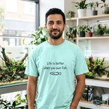 Man with a dark beard wearing the Life Is Better When You Own Fish tee in Chalky Mint, in an aquarium store in front of large planted display tanks