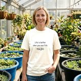 Woman with a blonde bob wearing the Life Is Better When You Own Fish tee in Ivory, in a greenhouse lined with tub ponds of water lettuce and frogbit