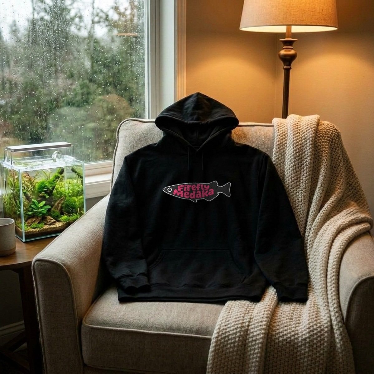 Firefly Medaka embroidered logo hoodie in black laid on a reading chair by a rainy window, next to a planted nano aquarium