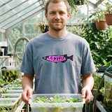 Man smiling in the Firefly Medaka logo heavyweight tee in Blue Jean, holding a tub of floating water lettuce in a greenhouse