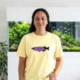 Woman with long dark wavy hair wearing the Firefly Medaka logo heavyweight tee in Butter, in a living room in front of a large planted aquarium