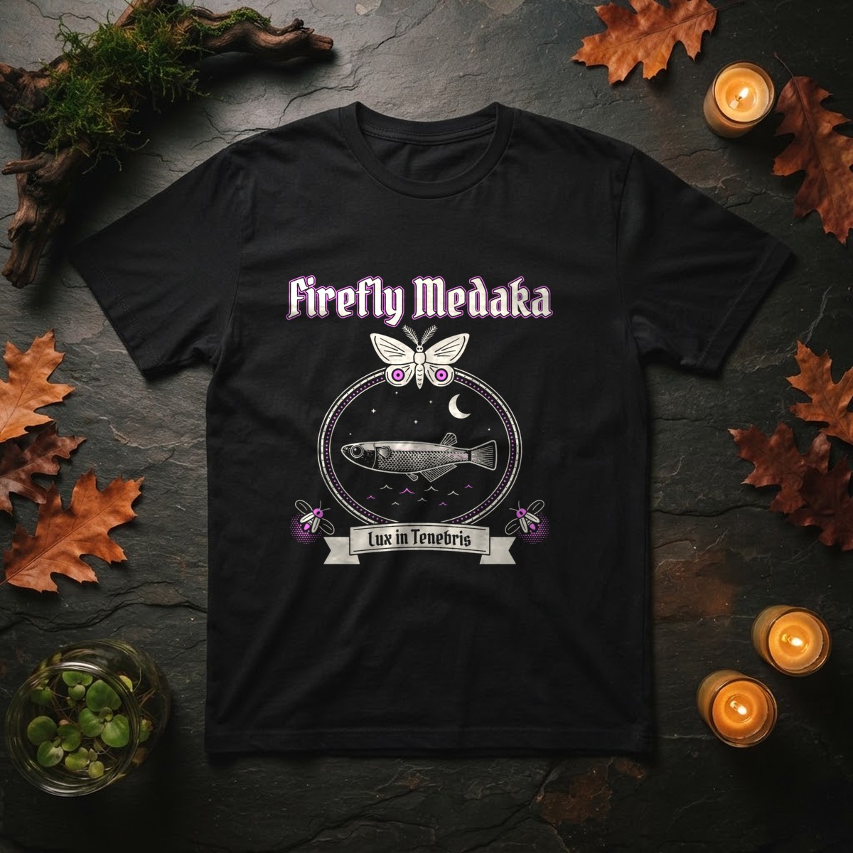 Lux in Tenebris goth medaka t-shirt in black, laid flat on dark slate with mossy aquarium driftwood, a jar of floating frogbit, autumn oak leaves, and lit candles