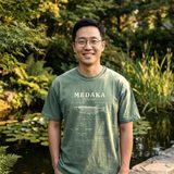Man with round glasses wearing the Medaka Field Guide tee in Blue Spruce, at the stone edge of a garden pond with lily pads