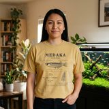Woman with long black hair wearing the Medaka Field Guide tee in Mustard, in a living room beside a large planted aquarium