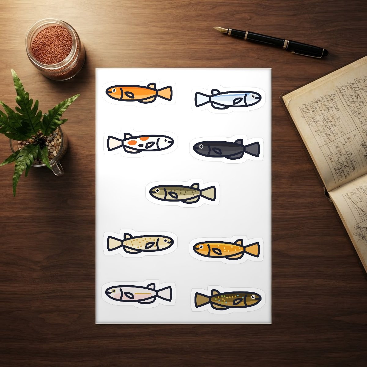 Medaka sticker sheet with nine kiss-cut rice fish stickers on a walnut desk beside a fish keeping logbook, a fountain pen, a jar of fish food, and a java fern