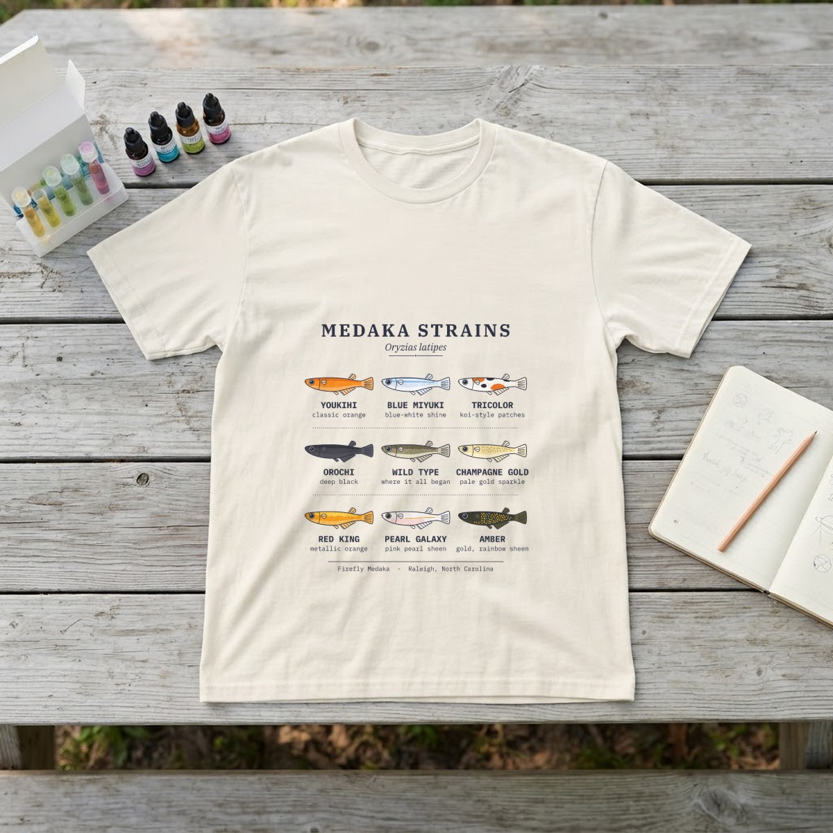 Medaka strains field chart t-shirt in ivory, laid flat on a weathered gray picnic table beside an aquarium water test kit, dropper bottles, and an open notebook