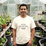 Man with a mustache wearing the Rice Fish, Not a Sushi Order tee in Ivory, in a greenhouse with large black tub ponds along the floor