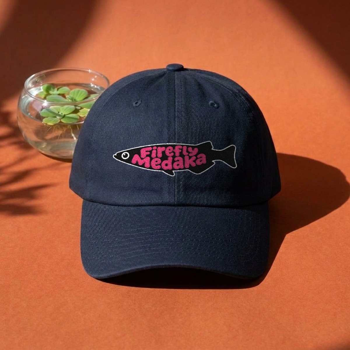 Firefly Medaka embroidered logo USA-made dad hat in navy on a terracotta backdrop beside a glass bowl of floating frogbit